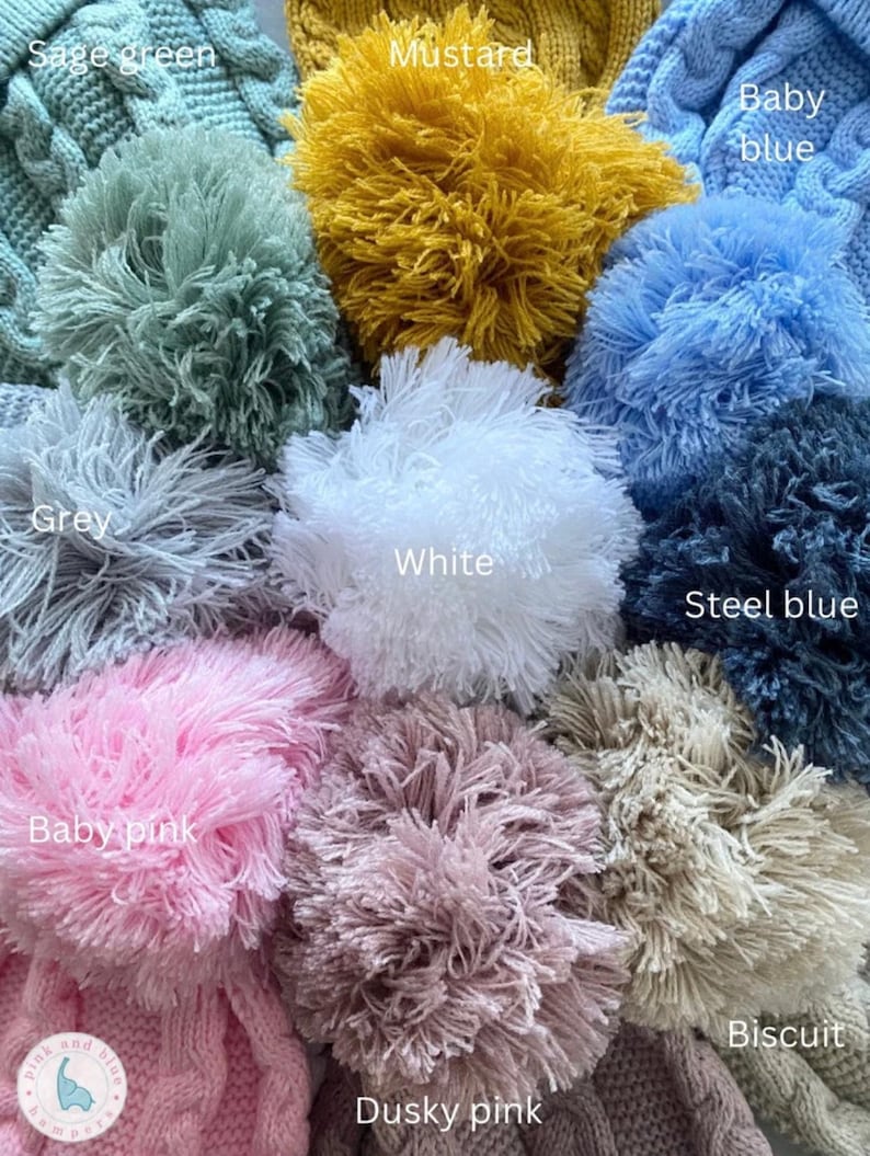 May include: A collection of knitted hats featuring large yarn pom-poms in various colors including sage green, mustard, baby blue, grey, white, steel blue, baby pink, dusky pink, and biscuit, with text labels identifying each color shade.