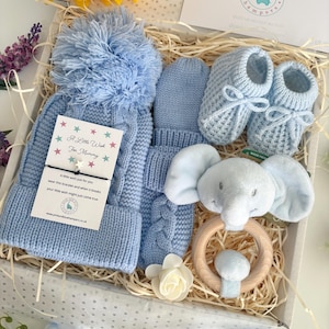May include: A baby gift set in a white box containing a light blue cable-knit hat with a pom-pom, matching mittens, knitted booties with bows, and a plush elephant rattle on a wooden ring. A card reads A Little Wish For Mummy.