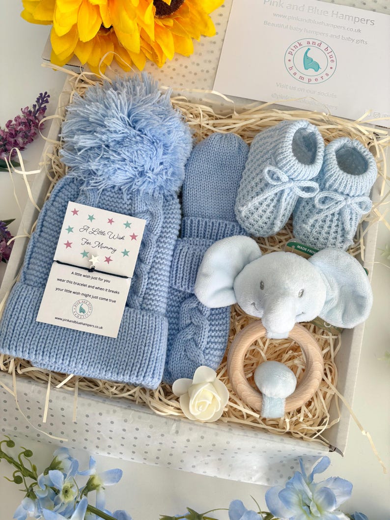 May include: A baby gift set in a white box containing a light blue cable-knit hat with a pom-pom, matching mittens, knitted booties with bows, and a plush elephant rattle on a wooden ring. A card reads A Little Wish For Mummy.
