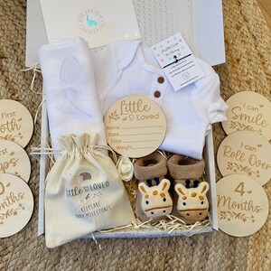 Pode incluir: Uma caixa de presente branca cheia de itens para beb&ecirc;s, incluindo um macac&atilde;o branco, um cobertor branco, uma pequena bolsa de juta com o texto "Little & Loved Keepsake Baby Milestones", uma placa de madeira que diz "Little & Loved" com espa&ccedil;os para preencher o nome do beb&ecirc;, a data, a hora e o peso, e um conjunto de discos de marcos de madeira com frases como "My First Tooth", "My First Steps", "10 Months", "I Can Smile", "I Can Roll Over" e "4 Months".