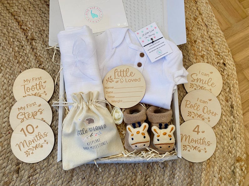 Pode incluir: Uma caixa de presente branca cheia de itens para beb&ecirc;s, incluindo um macac&atilde;o branco, um cobertor branco, uma pequena bolsa de juta com o texto "Little & Loved Keepsake Baby Milestones", uma placa de madeira que diz "Little & Loved" com espa&ccedil;os para preencher o nome do beb&ecirc;, a data, a hora e o peso, e um conjunto de discos de marcos de madeira com frases como "My First Tooth", "My First Steps", "10 Months", "I Can Smile", "I Can Roll Over" e "4 Months".