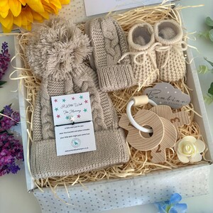 May include: A beige knit baby gift set in a box, including a cabled hat with a large pom-pom, matching mittens, and booties. It features a wooden key-shaped teether, a small white flower, and a card with the text A Little Wish For Mommy.