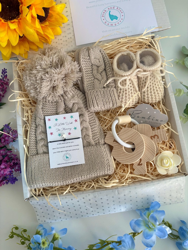 May include: A beige knit baby gift set in a box, including a cabled hat with a large pom-pom, matching mittens, and booties. It features a wooden key-shaped teether, a small white flower, and a card with the text A Little Wish For Mommy.