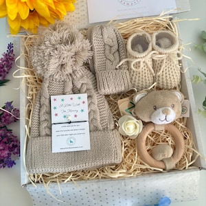 May include: A gift box containing a beige cable-knit hat with a pom-pom, matching mittens, and booties. It includes a wooden ring rattle with a plush bear head, a white artificial flower, and a card with the text A Little Wish For Mommy.