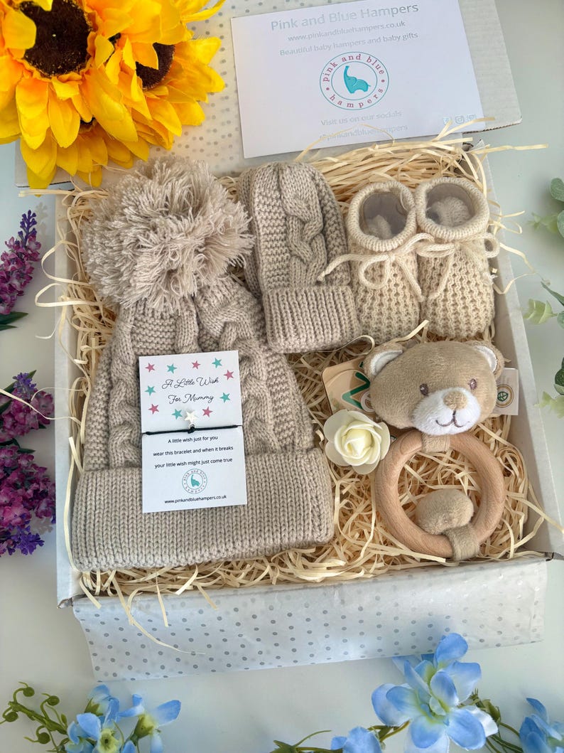 May include: A gift box containing a beige cable-knit hat with a pom-pom, matching mittens, and booties. It includes a wooden ring rattle with a plush bear head, a white artificial flower, and a card with the text A Little Wish For Mommy.