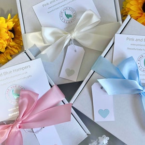 May include: Two white gift boxes with pink and blue ribbons and tags. The boxes are tied with bows and have a heart-shaped tag on each. The boxes are labeled "Pink and Blue Hampers" and have a website address and social media handle.