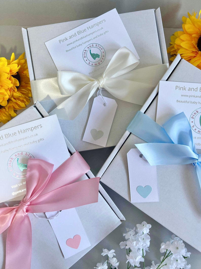 May include: Two white gift boxes with pink and blue ribbons and tags. The boxes are tied with bows and have a heart-shaped tag on each. The boxes are labeled "Pink and Blue Hampers" and have a website address and social media handle.