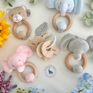May include: A collection of baby rattles featuring plush animal heads including a tan bear, a light blue elephant, a pink elephant, and a gray elephant, each attached to a wooden ring. Also included are wooden number charms and a circular logo tag.