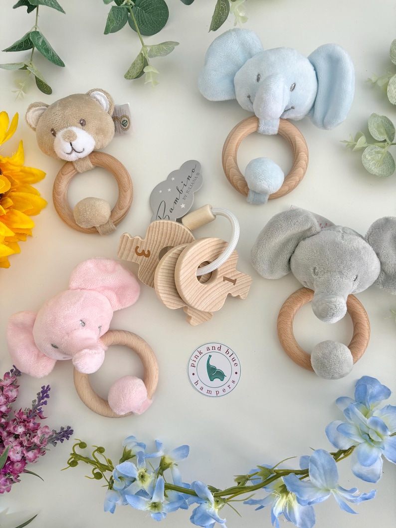 May include: A collection of baby rattles featuring plush animal heads including a tan bear, a light blue elephant, a pink elephant, and a gray elephant, each attached to a wooden ring. Also included are wooden number charms and a circular logo tag.