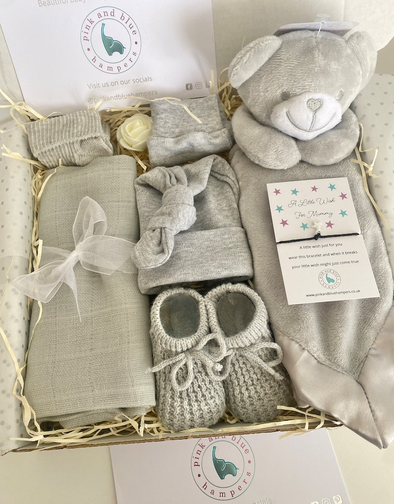 May include: A gray and white baby gift box with a gray knitted hat, booties, socks, a plush gray teddy bear, a gray blanket, and a white card with the text "A Little Wish For Mummy".