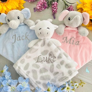 May include: Three personalized baby blankets with plush animal toys attached. The blankets are blue, pink, and white with gray spots. The toys are a gray elephant, a gray elephant, and a white cow. The blankets are embroidered with the names "Jack", "Mia", and "Luke".