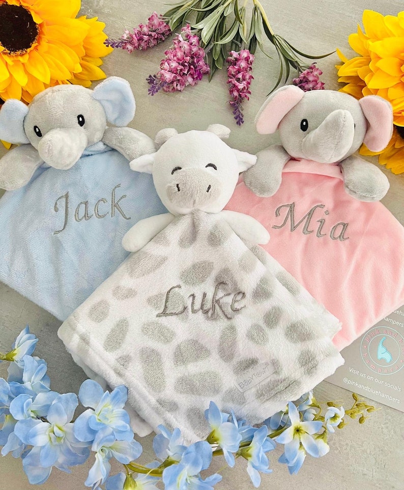 May include: Three personalized baby blankets with plush animal toys attached. The blankets are blue, pink, and white with gray spots. The toys are a gray elephant, a gray elephant, and a white cow. The blankets are embroidered with the names "Jack", "Mia", and "Luke".