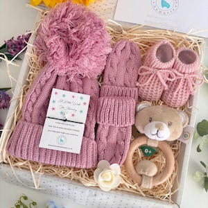 May include: A gift box containing a dusty pink cable-knit hat with a large pom-pom, matching cable-knit mittens, a pair of pink knitted booties, a plush teddy bear rattle with a wooden ring, and a card with a string bracelet labeled A Little Wish.