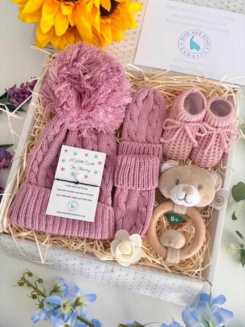 May include: A gift box containing a dusty pink cable-knit hat with a large pom-pom, matching cable-knit mittens, a pair of pink knitted booties, a plush teddy bear rattle with a wooden ring, and a card with a string bracelet labeled A Little Wish.