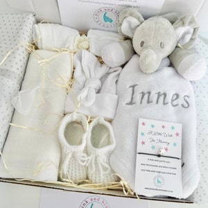 May include: A white gift box filled with baby items, including a white knitted baby hat, a white blanket, a pair of white knitted booties, a white elephant plush toy, and a white towel with the name "Innes" embroidered on it. The box also includes a small card with the text "A Little Wish For Mommy" and a white bracelet with a star charm.