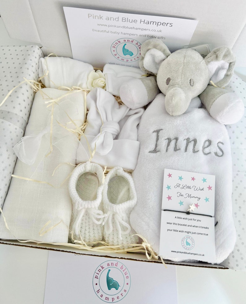 May include: A white gift box filled with baby items, including a white knitted baby hat, a white blanket, a pair of white knitted booties, a white elephant plush toy, and a white towel with the name "Innes" embroidered on it. The box also includes a small card with the text "A Little Wish For Mommy" and a white bracelet with a star charm.