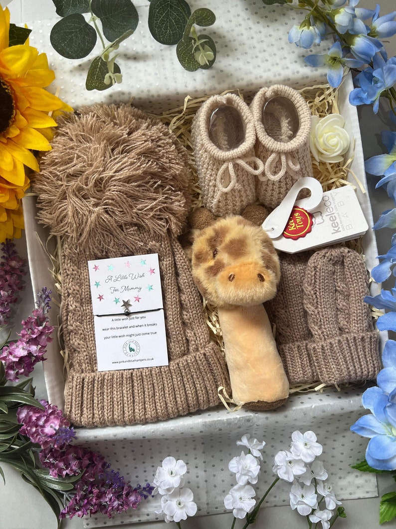 May include: A gift set containing a tan cable-knit baby hat with a large pom-pom, matching tan knit baby booties, tan knit mittens, and a plush giraffe rattle. A card with the text A Little Wish For Mummy and a small star charm is included.