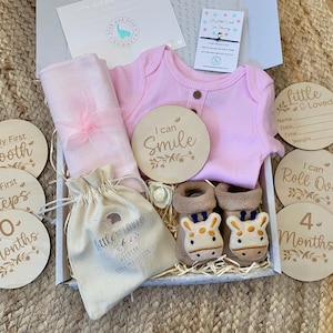 Pode incluir: Um macac&atilde;o rosa para beb&ecirc;, um envolt&oacute;rio de musselina rosa, uma pequena bolsa branca com o texto "Little & Loved Keepsake Baby Milestones", e um conjunto de discos de madeira para marcos de beb&ecirc; com frases como "Meu primeiro dente", "Eu posso sorrir", "Eu posso rolar", e "4 meses".