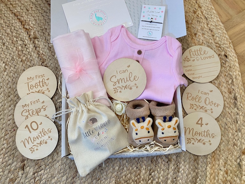 Pode incluir: Um macac&atilde;o rosa para beb&ecirc;, um envolt&oacute;rio de musselina rosa, uma pequena bolsa branca com o texto "Little & Loved Keepsake Baby Milestones", e um conjunto de discos de madeira para marcos de beb&ecirc; com frases como "Meu primeiro dente", "Eu posso sorrir", "Eu posso rolar", e "4 meses".