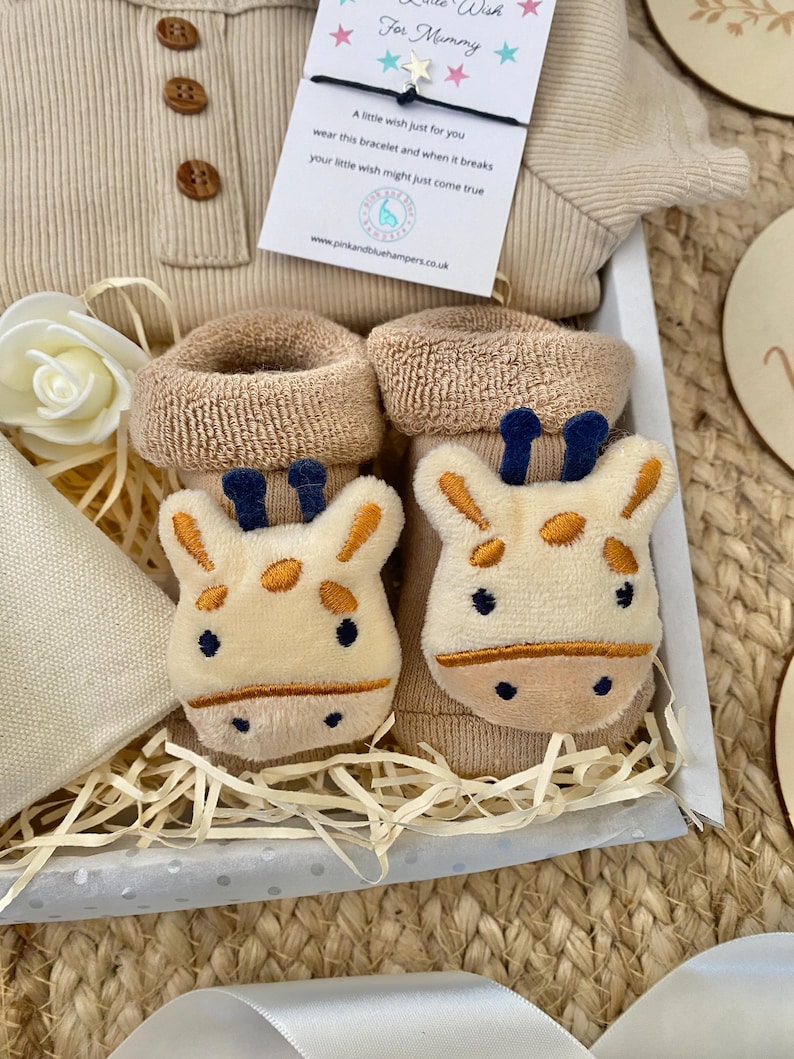 Pode incluir: Uma cesta de presente para um rec&eacute;m-nascido com um macac&atilde;o bege, um par de sapatinhos de beb&ecirc; bege com um rosto de girafa, um cobertor branco e bege e um cart&atilde;o pequeno com o texto "Pequeno desejo para mam&atilde;e".