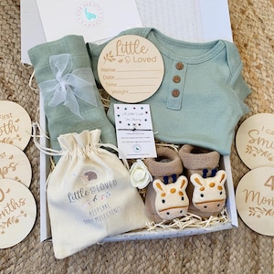 Pode incluir: Uma caixa de presente cheia de itens para beb&ecirc;s, incluindo um macac&atilde;o verde, um cobertor de musselina branco, uma pequena bolsa com o texto "Little & Loved Keepsake Baby Milestones" e placas de marcos de madeira com frases como "My First Tooth", "My First Steps", "10 Months", "I Can Smile" e "4 Months".