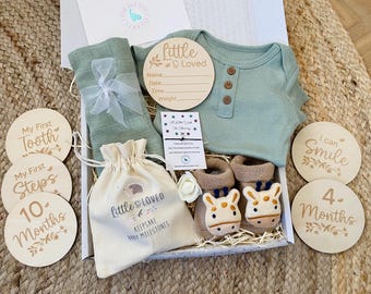 Può includere: Una scatola regalo piena di articoli per bambini, tra cui un body verde, una copertina in mussola bianca, una piccola borsa con il testo "Little & Loved Keepsake Baby Milestones" e targhe in legno con frasi come "My First Tooth", "My First Steps", "10 Months", "I Can Smile" e "4 Months".