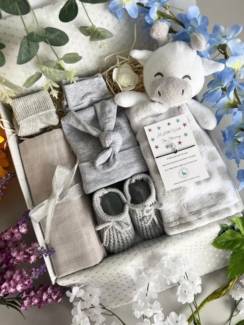 May include: A gray and white baby gift set in a white box with a white stuffed giraffe toy, a gray knitted hat, a gray knitted blanket, a gray knitted pair of booties, and a gray knitted sock. The box is decorated with white polka dots and the text "A Little Wish For Mummy".