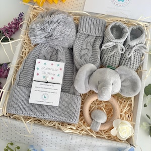May include: A gift box containing a gray knit hat with a pom-pom, gray knit mittens, gray knit booties with laces, and a gray plush elephant rattle on a wooden ring. A card with the text A Little Wish For Mummy is attached to the hat.