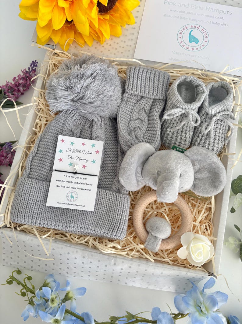 May include: A gift box containing a gray knit hat with a pom-pom, gray knit mittens, gray knit booties with laces, and a gray plush elephant rattle on a wooden ring. A card with the text A Little Wish For Mummy is attached to the hat.