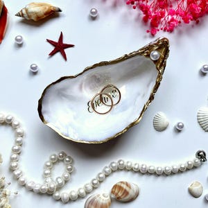 Könnte beinhalten: Eine weiße Austernschalenschale mit goldfarbenem Rand, die zwei goldene Ringe mit der Gravur Eveline enthält. Die Schale ist umgeben von einer weißen Perlenkette, verschiedenen kleinen Muscheln, einem roten Seestern und rosa Trockenblumen.