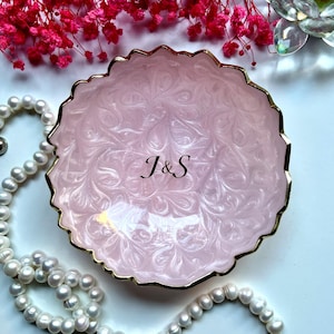 May include: A round, scalloped-edge trinket dish in a pale pink pearlescent finish with a gold-toned rim. The center of the dish features the black printed letters J&S in a serif font. The dish has a swirling, marbled texture throughout.