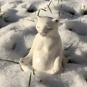 Könnte beinhalten: Eine weiße Keramik-Katzenfigur in einer sitzenden Yoga-Pose mit geschlossenen Augen. Die Katze befindet sich im Schnee, mit sichtbaren Grashalmen. Die Figur hat eine glatte, glänzende Oberfläche.