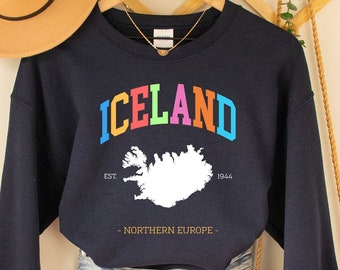 Puede incluir: Sudadera azul marino con la palabra "ICELAND" en letras coloridas. Debajo, un contorno blanco de Islandia y el texto "EST. 1944 - NORTHERN EUROPE". La sudadera se combina con pantalones cortos de mezclilla y un sombrero beige.