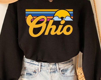 May include: A black long-sleeve sweatshirt featuring a graphic print of the word Ohio in yellow cursive script, positioned below a horizontal sunset design with blue, orange, and yellow stripes and a yellow sun rising over blue clouds.