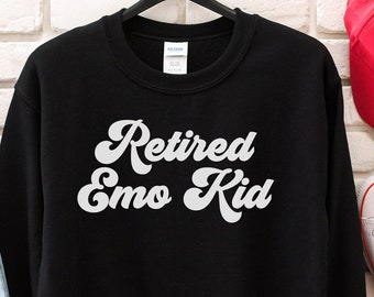 Könnte beinhalten: Ein schwarzer Rundhals-Pullover mit dem weißen Schriftzug Retired Emo Kid in einer Retro-Schreibschrift. Das Kleidungsstück hängt an einem schwarzen Kleiderbügel, ergänzt durch eine hellblaue Jeans im Used-Look, eine rote Kappe und weiße Sneaker.