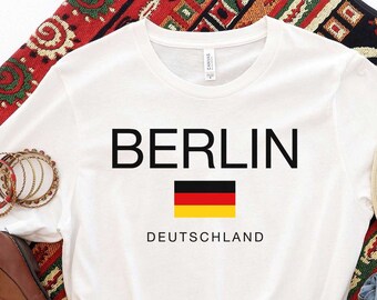 Puede incluir: Camiseta blanca con la palabra "BERLIN" en negro, una bandera alemana y "DEUTSCHLAND" debajo. La camiseta se combina con botas marrones, un sombrero de paja, pantalones cortos de mezclilla y pulseras doradas sobre una alfombra estampada.