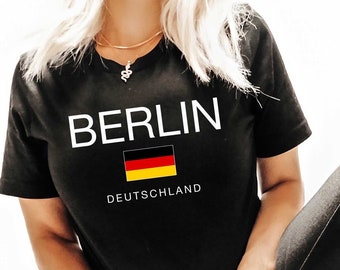 Puede incluir: Camiseta negra con la palabra "BERLIN" en blanco, una bandera alemana y la palabra "DEUTSCHLAND" en blanco. La persona lleva pantalones de cuero negros y botas de combate negras.