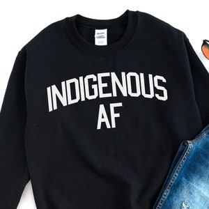 Può includere: Felpa nera con testo bianco che recita "Indigenous AF". La felpa è stesa su una superficie bianca con un paio di jeans blu, un paio di scarpe da ginnastica rosse e alcune foglie autunnali.
