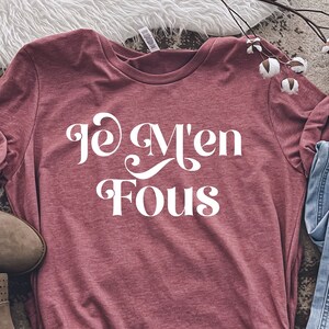 Puede incluir: Una camiseta de color marrón rojizo con el texto "Je M'en Fous" en letras blancas. La camiseta está colocada sobre una superficie con un patrón marrón y blanco, junto a un par de botines marrones de tobillo y unos vaqueros azules.
