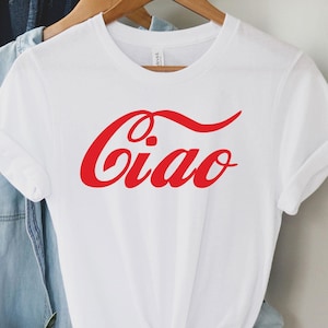 Puede incluir: Camiseta blanca con un logotipo rojo estilo Coca-Cola que dice "Ciao" sobre un fondo blanco.