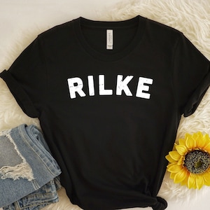 May include: Black t-shirt with white text that reads "RILKE" on a white background. The shirt is folded and laid on a white fluffy surface with a yellow sunflower.