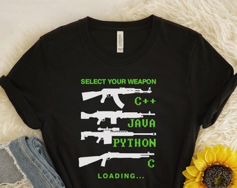 May include: Black t-shirt featuring the text "SELECT YOUR WEAPON" and gun illustrations, with programming languages C++, JAVA, PYTHON, and C listed below. The word "LOADING..." is at the bottom. The shirt is next to a pair of blue jeans and a yellow sunflower.