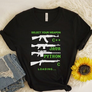 May include: Black t-shirt with the text "SELECT YOUR WEAPON" and images of guns, with the programming languages C++, JAVA, PYTHON, and C listed below. The word "LOADING..." is at the bottom. The shirt is next to a pair of blue jeans and a yellow sunflower.