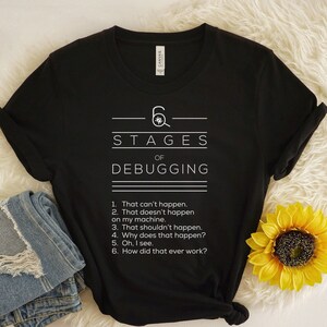以下が含まれることがあります： 白文字で「6 Stages of Debugging」と書かれた黒いTシャツ。青いジーンズと黄色のヒマワリが組み合わされています。テキストは、疑いから理解に至るまで、デバッグの段階をリストアップしています。