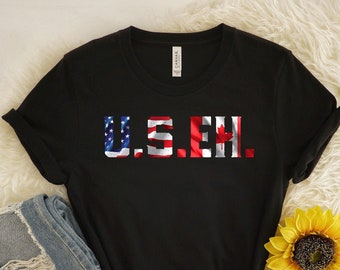 May include: Black t-shirt with the text "U.S. & CH." in a patriotic design, incorporating the American and Canadian flags. The shirt is paired with folded blue jeans and a yellow sunflower on a white fluffy surface.