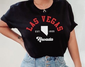 May include: A black short-sleeved t-shirt featuring the red text LAS VEGAS, a white silhouette of the state of Nevada, the text EST. 1905, and the word Nevada in white script. The shirt is paired with light blue denim jeans with a button-up fly.