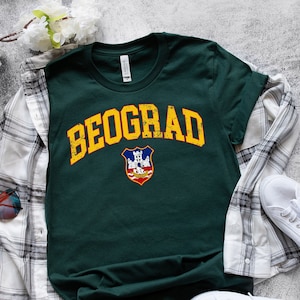 Puede incluir: Una camiseta de manga corta de color verde oscuro con el texto amarillo desgastado BEOGRAD en una fuente arqueada sobre un emblema de escudo colorido. La camiseta se combina con una camisa de cuadros blancos y negros, zapatillas y gafas.