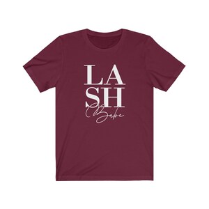 Pu&ograve; includere: T-shirt bordeaux a maniche corte con la scritta bianca &laquo;LASH Babe&raquo;. La parola &laquo;LASH&raquo; &egrave; impilata sopra &laquo;Babe&raquo;, con &laquo;SH&raquo; in mezzo. La parola &laquo;Babe&raquo; &egrave; in corsivo.