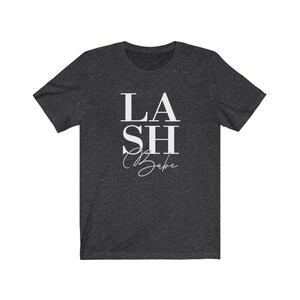 Pu&ograve; includere: T-shirt grigio scuro con la scritta bianca "LASH Babe". La parola "LASH" &egrave; in grassetto, con un carattere sans-serif, mentre "Babe" &egrave; in corsivo. La maglietta ha maniche corte e girocollo.