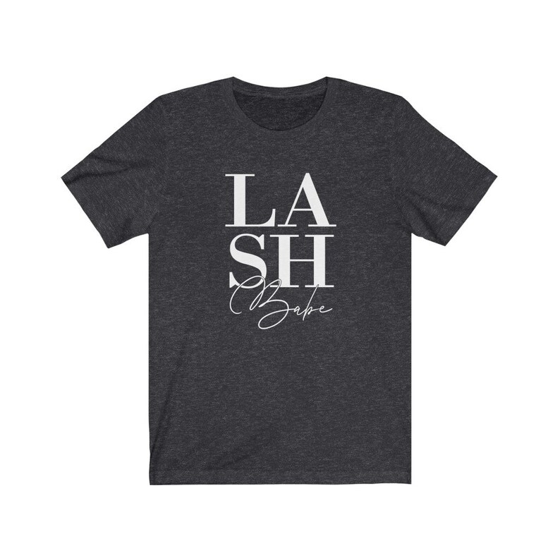 Pu&ograve; includere: T-shirt grigio scuro con la scritta bianca "LASH Babe". La parola "LASH" &egrave; in grassetto, con un carattere sans-serif, mentre "Babe" &egrave; in corsivo. La maglietta ha maniche corte e girocollo.
